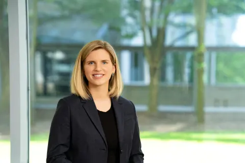 A woman in a dark blazer beside windows overlooking a courtyard.