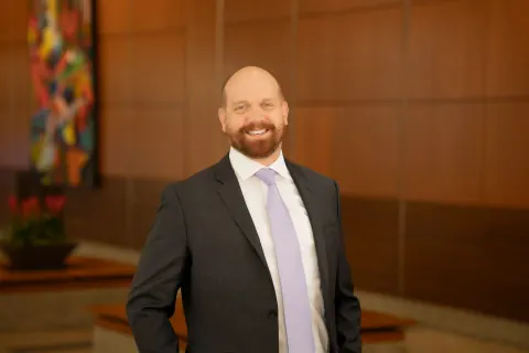 A smiling bearded man in a dark suit and pale tie against wood panelling.