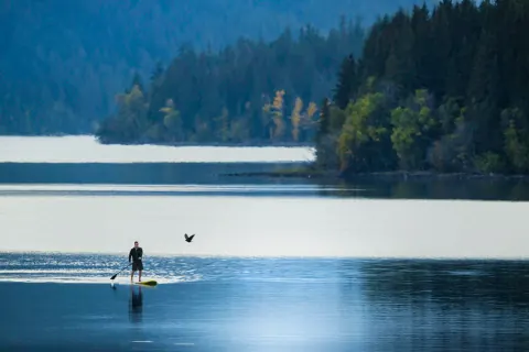 A paddleboarder on a still lake bordered by forested hills.