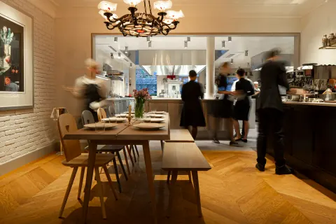 A bright restaurant interior with tables, a chandelier and staff at the bar.