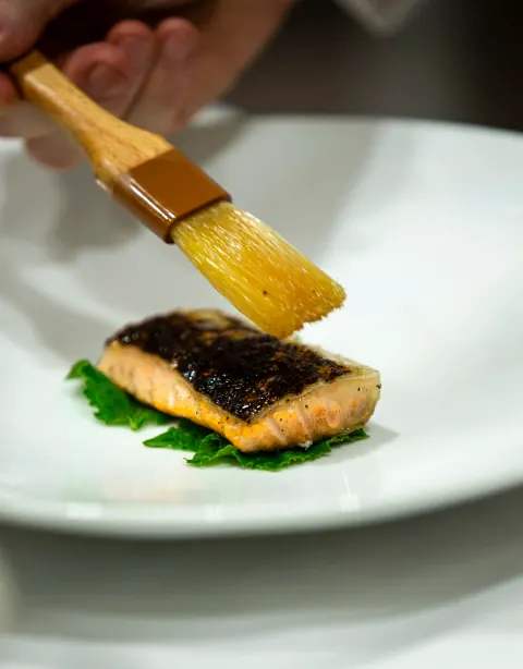 A pastry brush glazing a cooked fillet on a white plate.