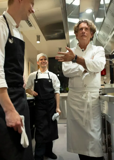 A chef speaking to staff in a stainless-steel kitchen.