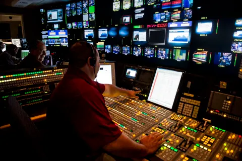 A person operating an illuminated audio mixing desk beneath rows of screens.