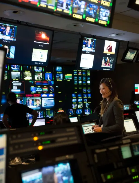 A woman at a broadcast control desk beneath multiple screens.