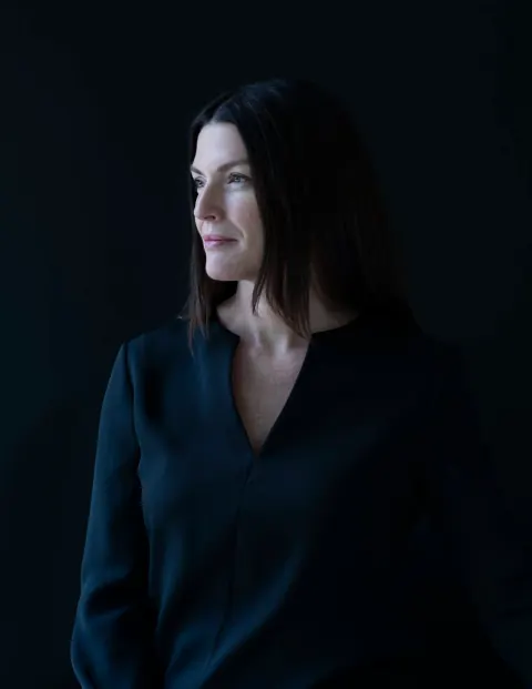 A woman in a dark blouse looking to the side against a dark backdrop.