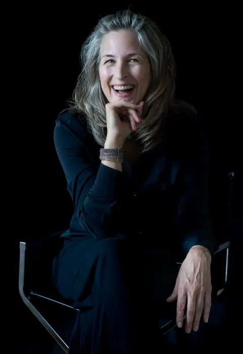 A smiling seated woman in black with her hand near her chin.