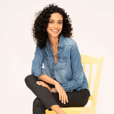 A smiling woman in a denim shirt seated on a yellow chair.