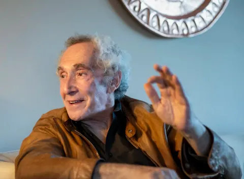 A grey-haired man gesturing with one hand while seated indoors.