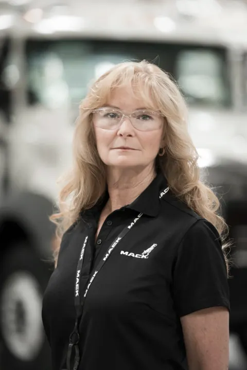 A woman in glasses and a black polo shirt against a blurred factory background.