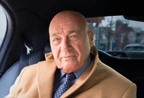 A bald man in a tan coat seated inside a car.