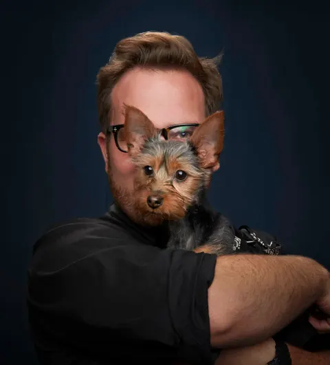 A man in glasses holding a small dog close to his face.