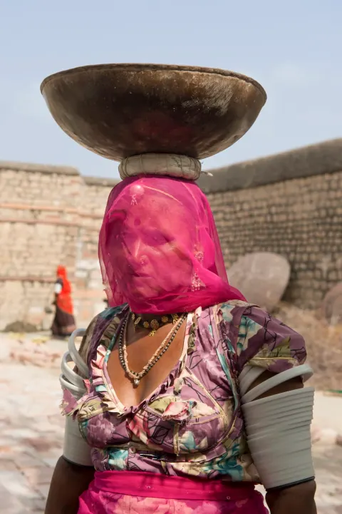 A woman wearing a pink veil and balancing a large bowl on her head.