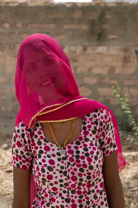A woman in a pink veil and spotted blouse against a brick wall.