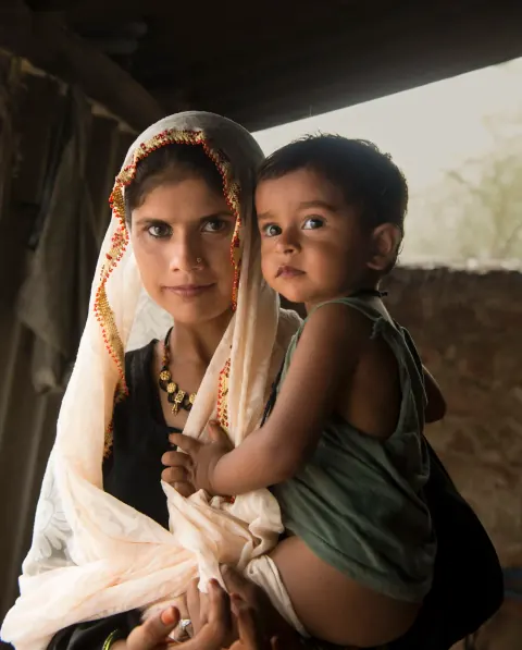 A woman in a pale scarf holding a child inside a shaded building.