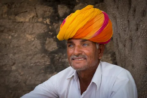 A smiling man in a yellow and orange turban beside a stone wall.