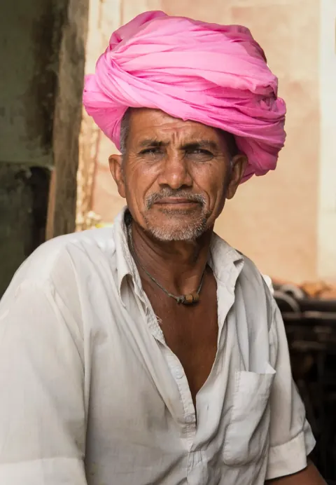 A man in a pink turban and white shirt seated by a doorway.