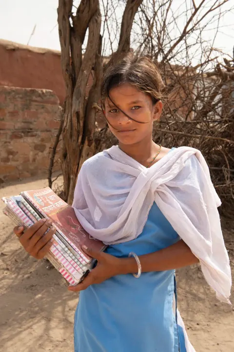 A girl in blue and white clothing holding a book beside a tree.
