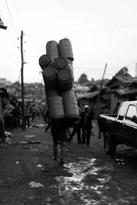 A monochrome street scene with a person carrying a tall stack of cylindrical objects.