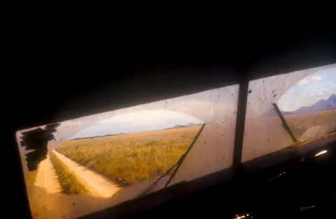 A sunlit dirt track seen through the dark windows of a vehicle.