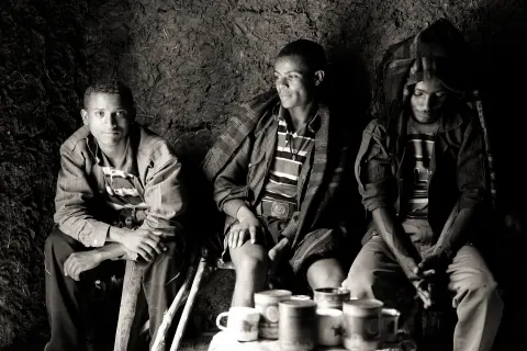 Three people seated around cups in a dark interior, photographed in black and white.